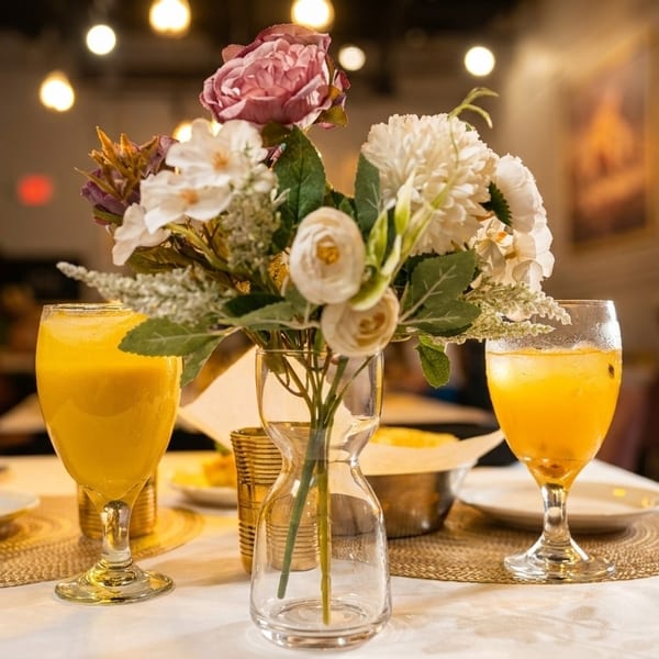 A restaurant table set with two drinks, plates, and a floral centerpiece, with a blurred background.