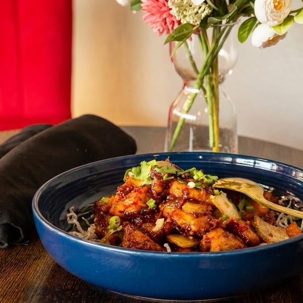 A blue bowl of General Tso's chicken with a gold spoon, black napkin, and flowers.
