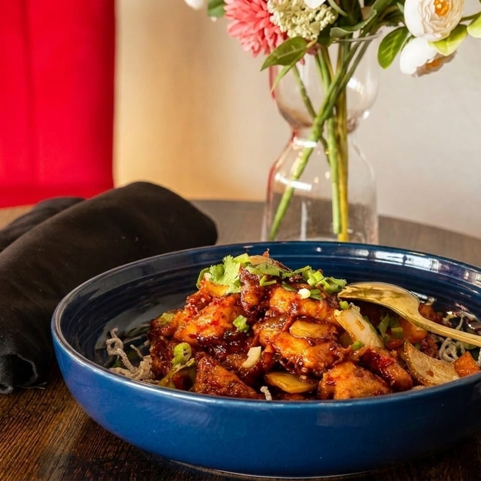 A blue bowl of General Tso's chicken with a gold spoon, black napkin, and flowers.