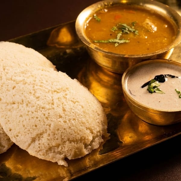 Three fluffy idlis with sambar, coconut chutney, and tomato chutney in brass bowls on a golden tray.